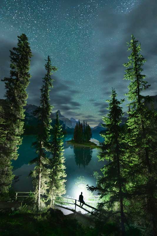 Spirit Island at night on Maligne Lake in Jasper National Park with Nick Fitzhardinge's silhouette sitting on a railing and illuminated by light.